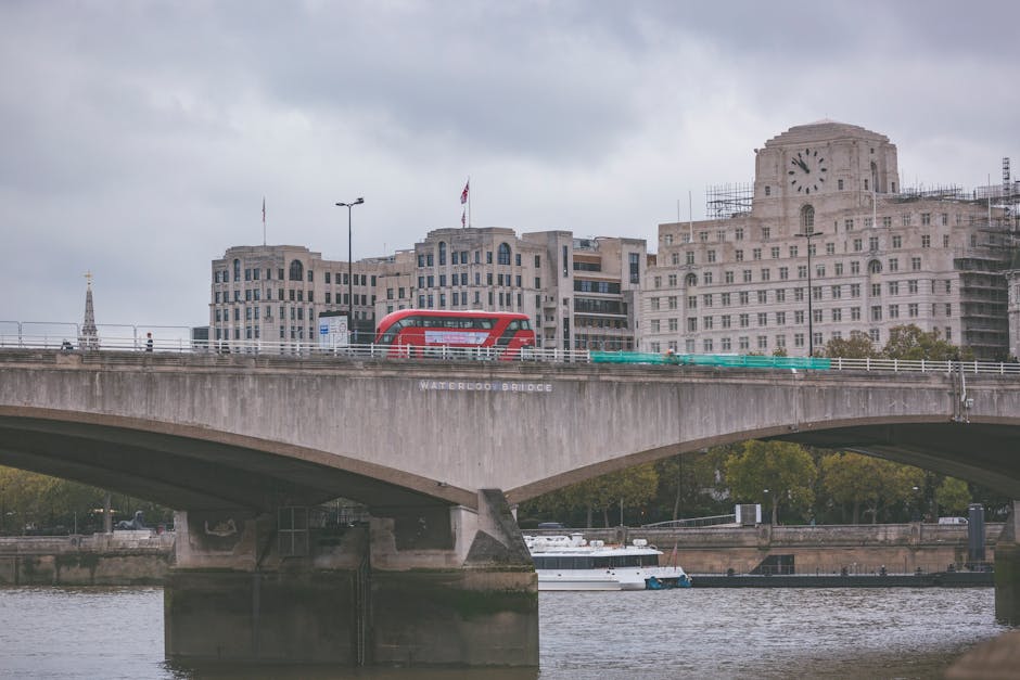 A panoramic view of Waterloo Bridge crossing the River Thames in London, with the bridge's concrete structure and arch visible in the foreground. On top of the bridge, a red double-decker bus is parked, and there are movement and activities suggesting loading or unloading during a home relocation process. Behind the bridge, there are several multi-storey buildings, including one with scaffolding and a large clock tower, indicating ongoing construction or renovation work. The sky above is overcast with grey clouds, creating a muted atmosphere. Downward, the river features a small white boat tied along the bank, while the area underneath the bridge appears calm. This scene captures the urban environment linked to shipping and transport logistics, relevant to house removals and furniture transport services provided by Man with Van Waterloo, situated near York Road flats, Waterloo.