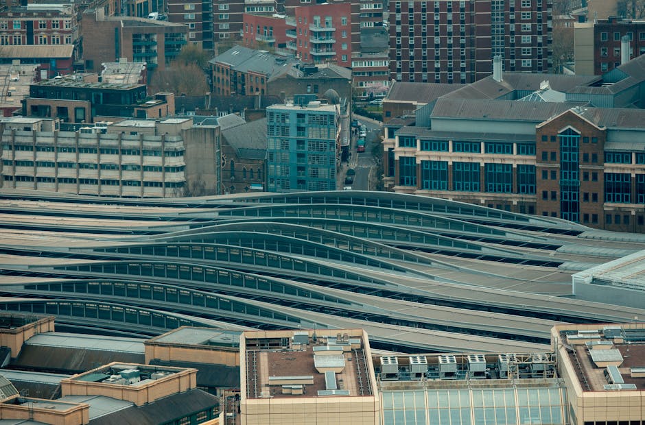 A high-angle view of a cityscape showing multiple modern, curving rooftop structures of a large transportation hub, likely Waterloo Station, with a background of densely packed office and residential buildings made of brick, glass, and concrete. The rooftop features elongated, wave-like metallic canopies with translucent panels, designed for shelter and light diffusion, arranged in parallel rows. Adjacent to the station, there are various adjacent rooftops, some with mechanical equipment and ventilation units. The image captures the urban environment during daytime with natural light illuminating the buildings, and the overall scene represents a busy metropolitan area, relevant to house removals and furniture transport focused on the Waterloo area, as indicated on the company website manwithvanwaterloo.co.uk.