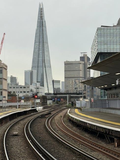 A panoramic view of Waterloo Bridge crossing the River Thames in London, with the bridge's concrete structure and arch visible in the foreground. On top of the bridge, a red double-decker bus is parked, and there are movement and activities suggesting loading or unloading during a home relocation process. Behind the bridge, there are several multi-storey buildings, including one with scaffolding and a large clock tower, indicating ongoing construction or renovation work. The sky above is overcast with grey clouds, creating a muted atmosphere. Downward, the river features a small white boat tied along the bank, while the area underneath the bridge appears calm. This scene captures the urban environment linked to shipping and transport logistics, relevant to house removals and furniture transport services provided by Man with Van Waterloo, situated near York Road flats, Waterloo.
